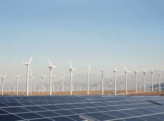 Field of wind turbines with solar panels in the foreground and mountains in the background.