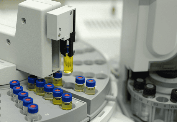 Laboratory machine handling vials with yellow liquid, some vials have blue caps and are arranged in a circular tray.