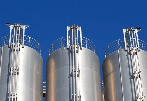 Three large stainless steel industrial silos with safety railings against a clear blue sky.