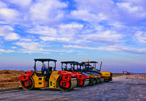 Road rollers and construction equipment on a dirt road under a partly cloudy sky.