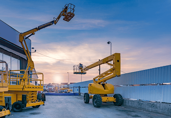 Two yellow boom lifts in use at a construction site during sunset.