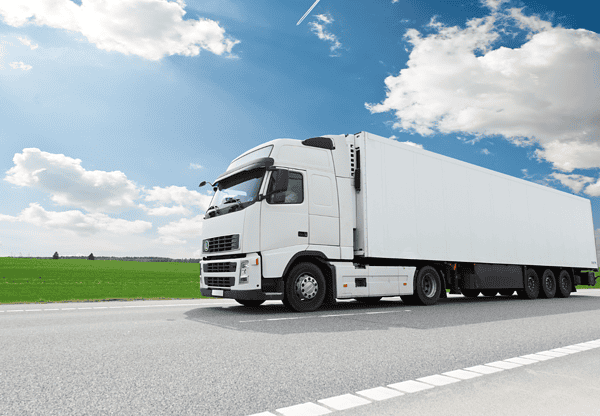 White semi-truck driving on a highway with green fields and blue sky in the background.