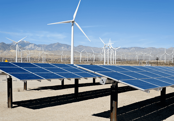 Solar panels and wind turbines in a desert landscape with mountains in the background.