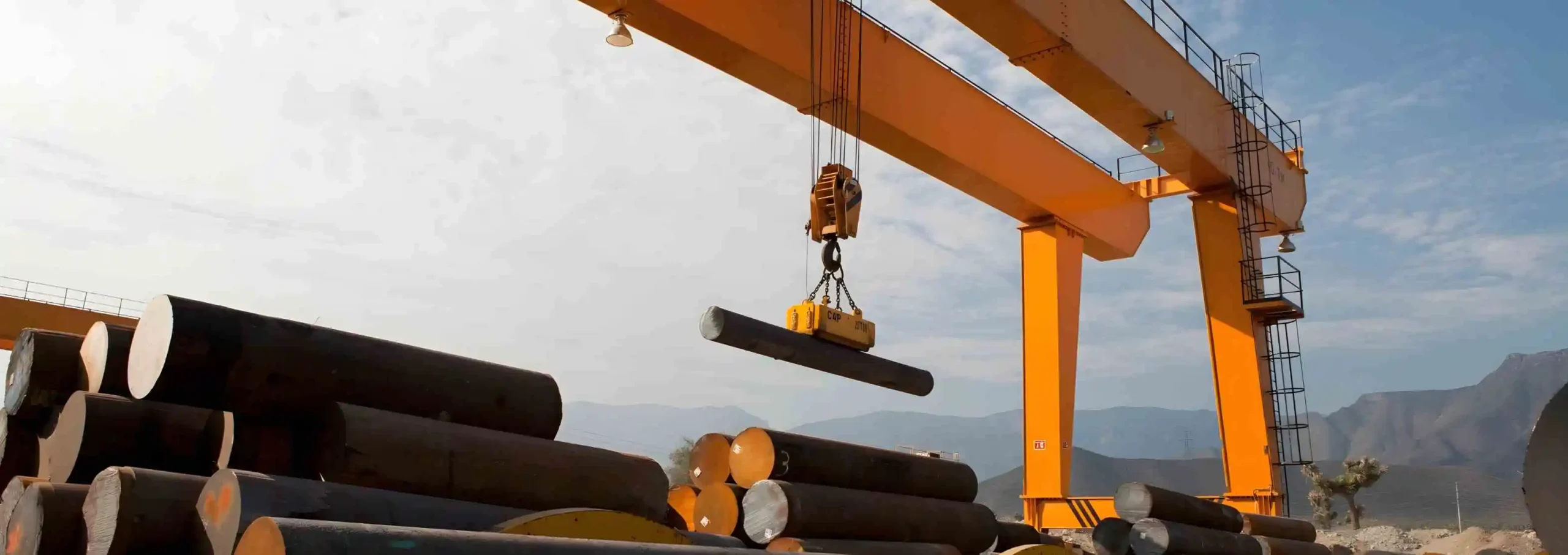 A large crane lifts an alloy steel bar in an industrial yard, with stacks of similar bars in the background.