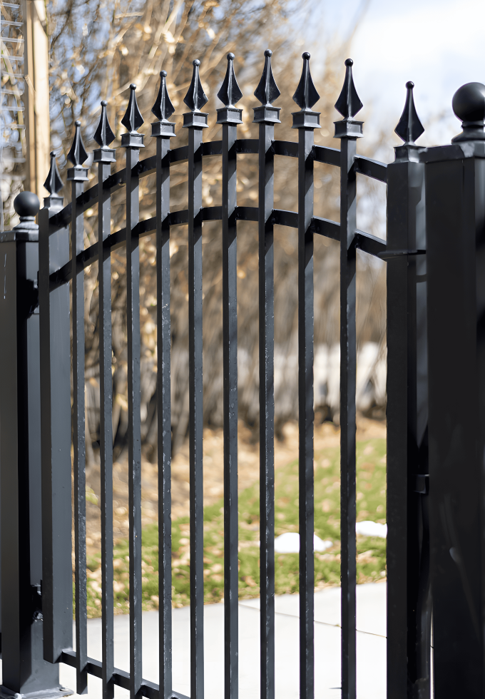 The photo shows a black wrought iron gate with pointed finials on top, situated outdoors with a backdrop of trees and a partly cloudy sky.