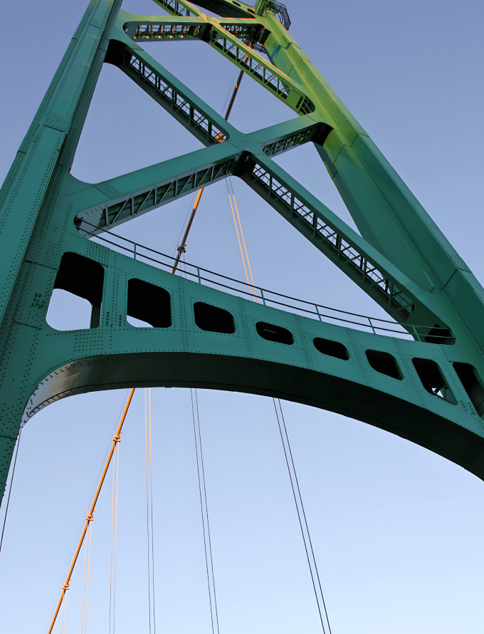 A close-up view of a green structural steel bridge with cables and a lamp post in the background.