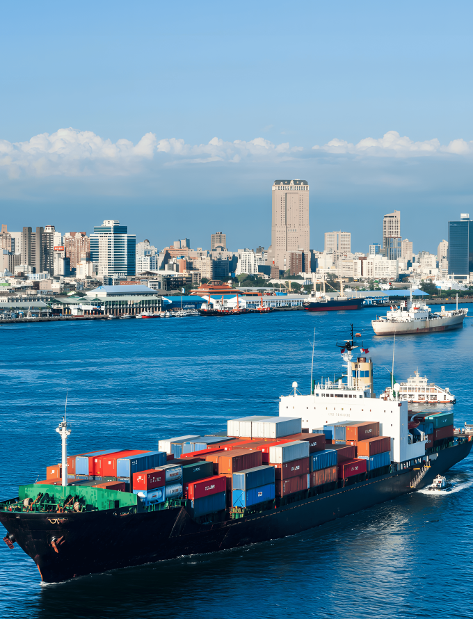 A cargo ship loaded with containers sailing in a harbor with a cityscape in the background.