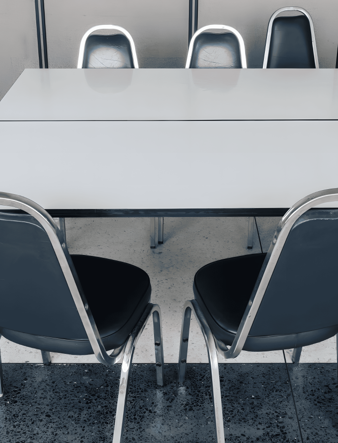 A white table surrounded by six blue chairs with metal frames, placed in a room with gray walls.