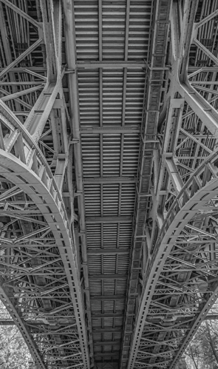 Black and white image of the underside of a metal bridge, showcasing its intricate structural design.