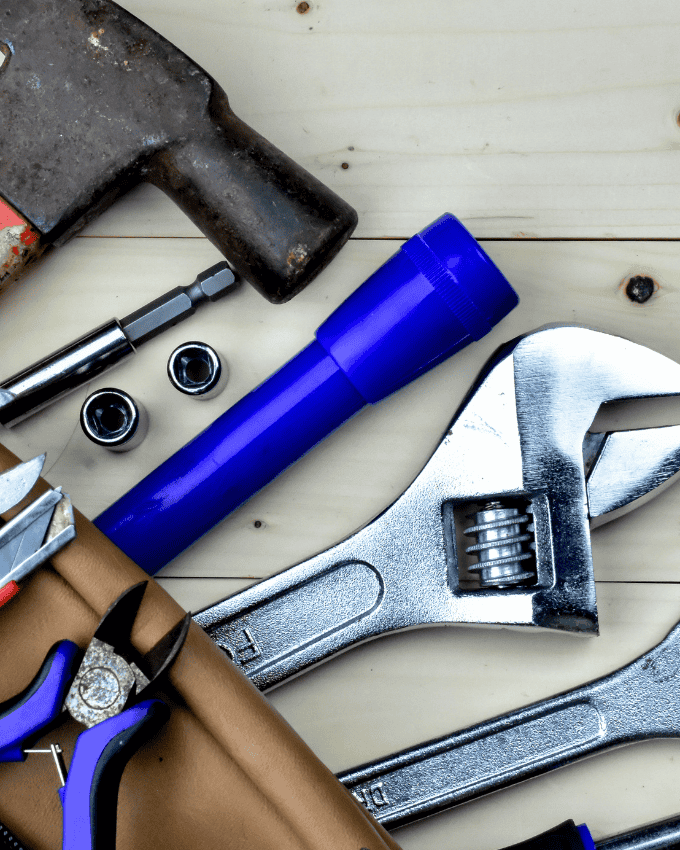 Assorted tools on a wooden surface, including a hammer, flashlight, wrench, pliers, and socket bits.