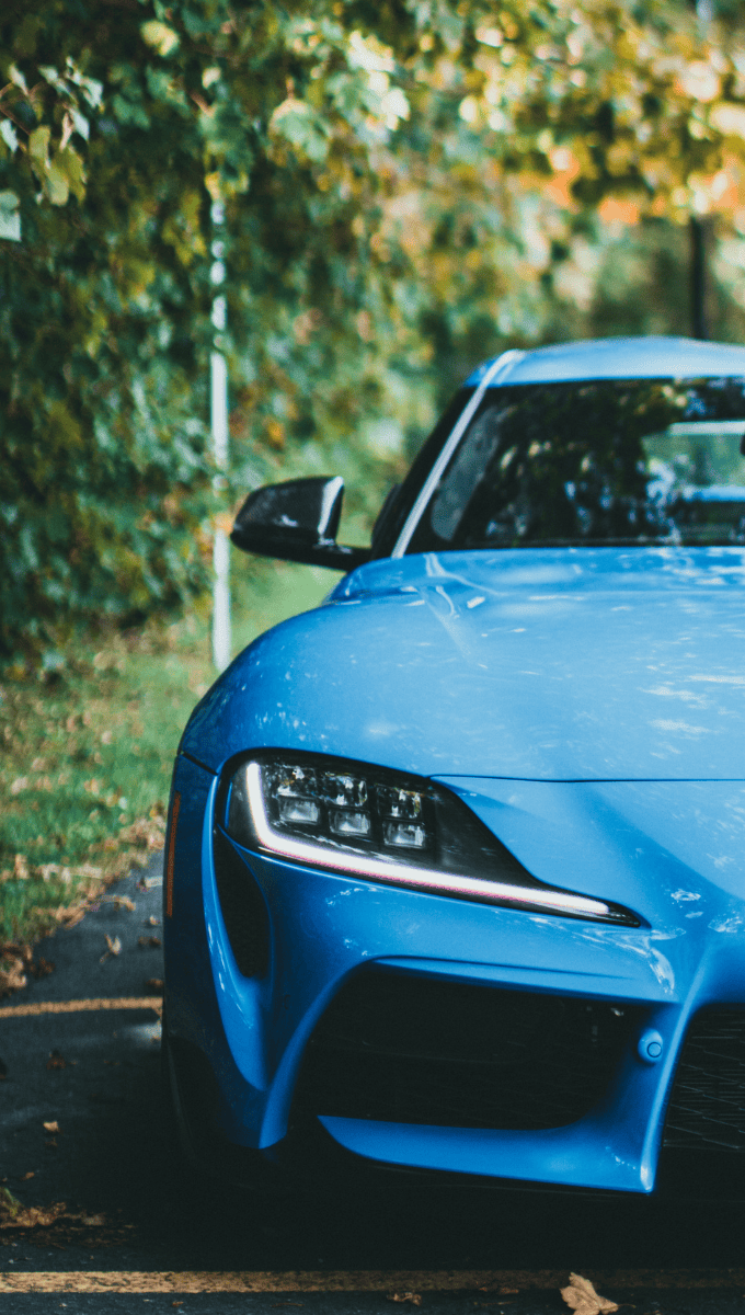 Vista frontal de un coche deportivo azul aparcado en una carretera con follaje verde de fondo.