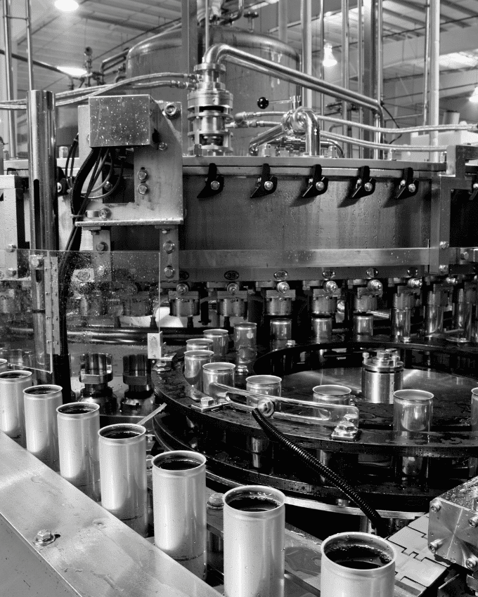 Imagen en blanco y negro de una máquina industrial en el entorno de una fábrica, con contenedores metálicos siendo procesados.