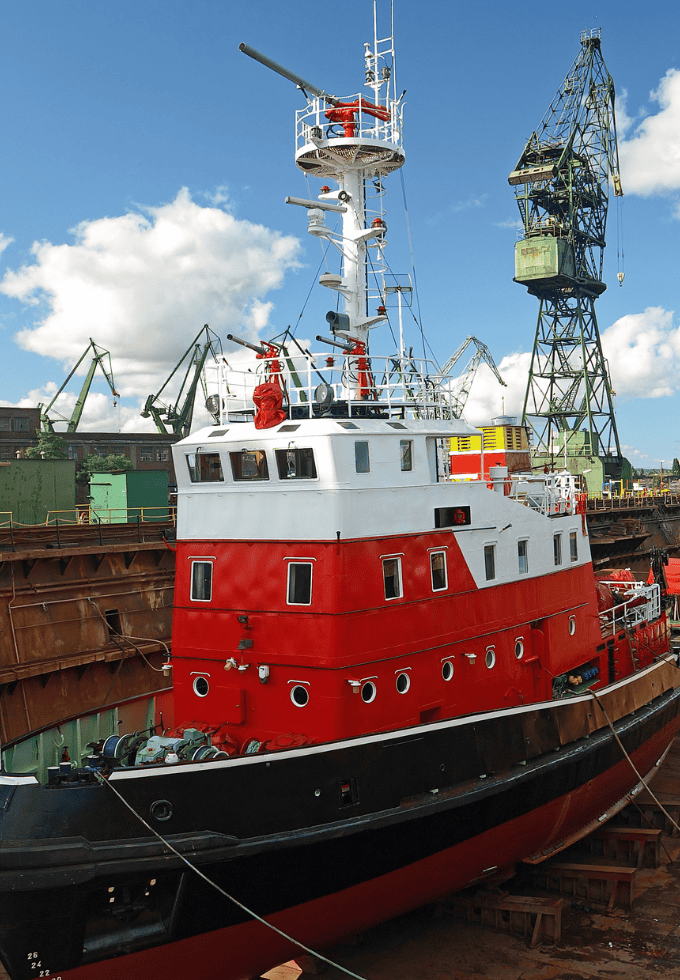 Red and white ship docked in a shipyard with cranes in the background.